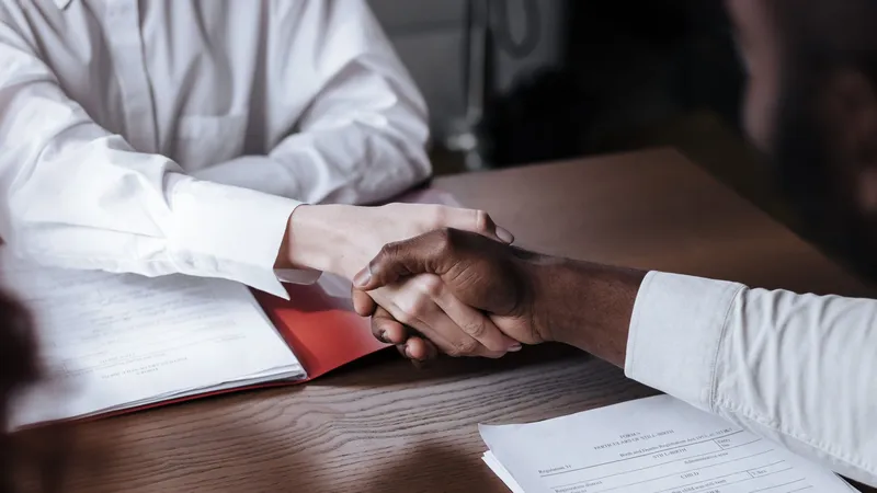Two people shaking hands across a desk covered in contract papers